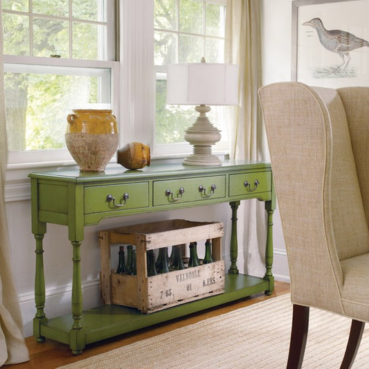 Green console table with a lamp and decorative items in a room with a window and chair.